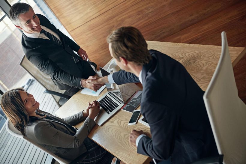 Two men shaking hands across a table while a woman in business attire smiles nearby.