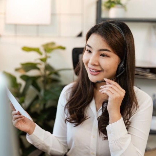 Smiling woman wearing headset holds paper while talking in an office setting with plants.